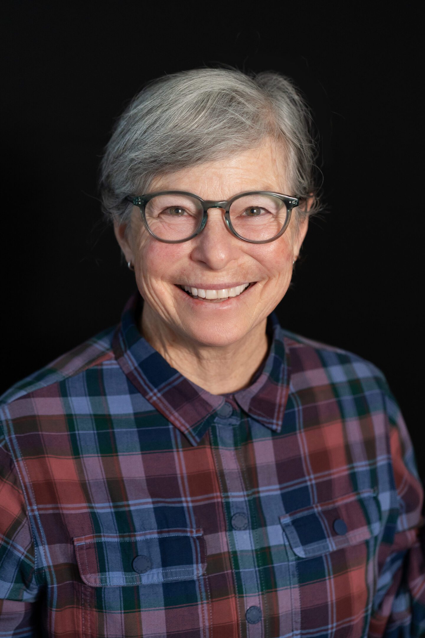 Smiling elderly woman in glasses and plaid shirt against black background.